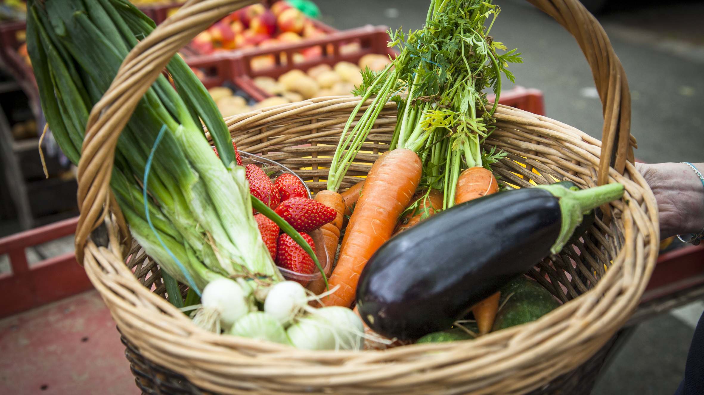 Panier de légumes en osier contenant des poireaux, des fraises, des carottes et des aubergines. Étal de pommes sur le marché en arrière-plan.